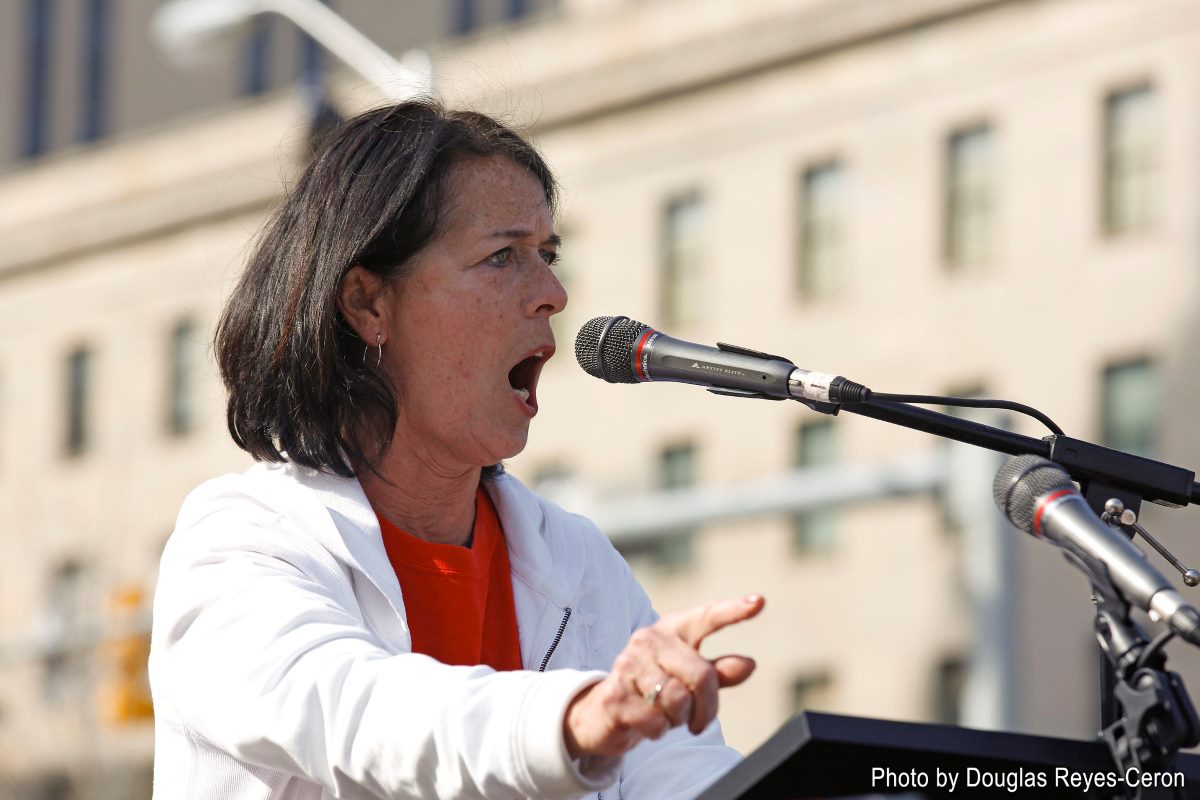 A woman in a white jacket speaks into a microphone at a podium, outdoors with a building in the background