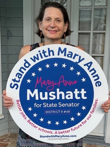 A woman stands holding a sign reading: "Stand with Mary Anne: Mary Anne Mushatt for State Senator."