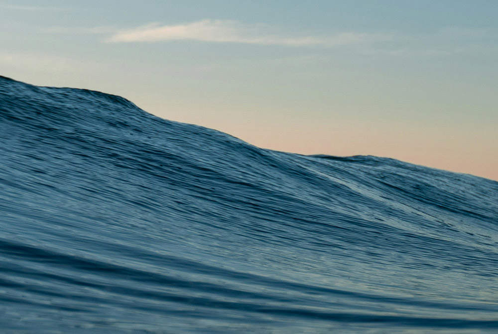 Blue wave in the ocean with sunset colors behind it.