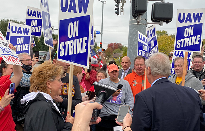 UAW workers at a press conference with signs showing they are on strike.