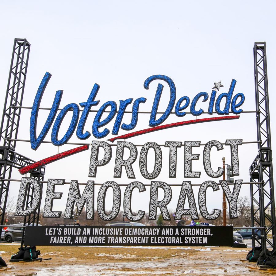 An art installation reads "Voters Decide, Protect our Democracy: Let's build an inclusive democracy and a stronger, fairer, and more transparent electoral system." The Capitol is seen in the background.