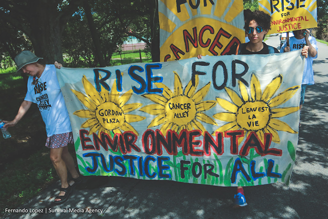 Two activists carry a colorful sign outdoors that reads, “Rise for Environmental Justice for All” and lists the examples of Gordon Plaza and Cancer Alley.